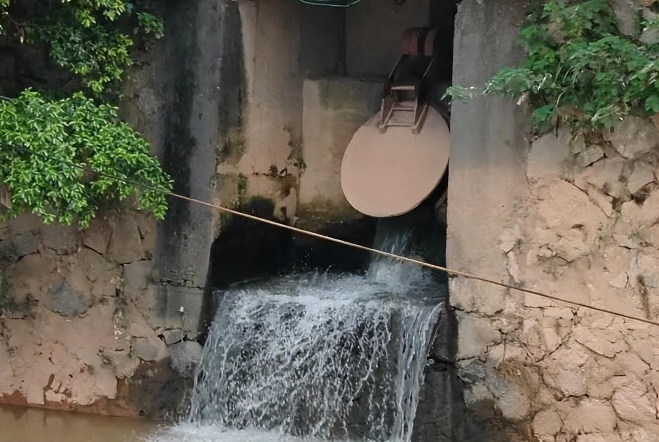石砌排水口闸门开启,水流从通道中倾泻而下,两侧绿植点缀斑驳石壁。